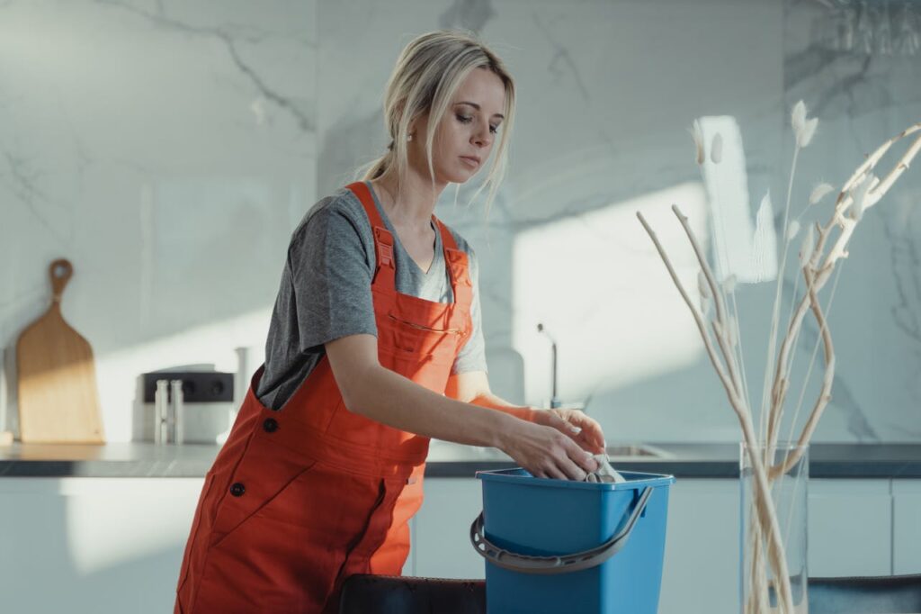 pexels-photo-6195111 A woman in a gray shirt and red overalls cleans a kitchen counter using a blue bucket and white rag. Modern setting.